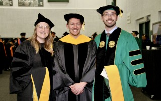 Three scientists stand next to eachother in graduation robes.